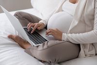 Schwangere sitzt mit Laptop auf dem Bett
