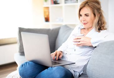Ältere Dame sitzt gemütlich auf der Couch und schaut in ihren Laptop
